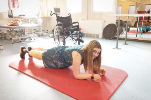 Vicky doing a plank on a red mat in a nurse's office with a wheelchair in the back.